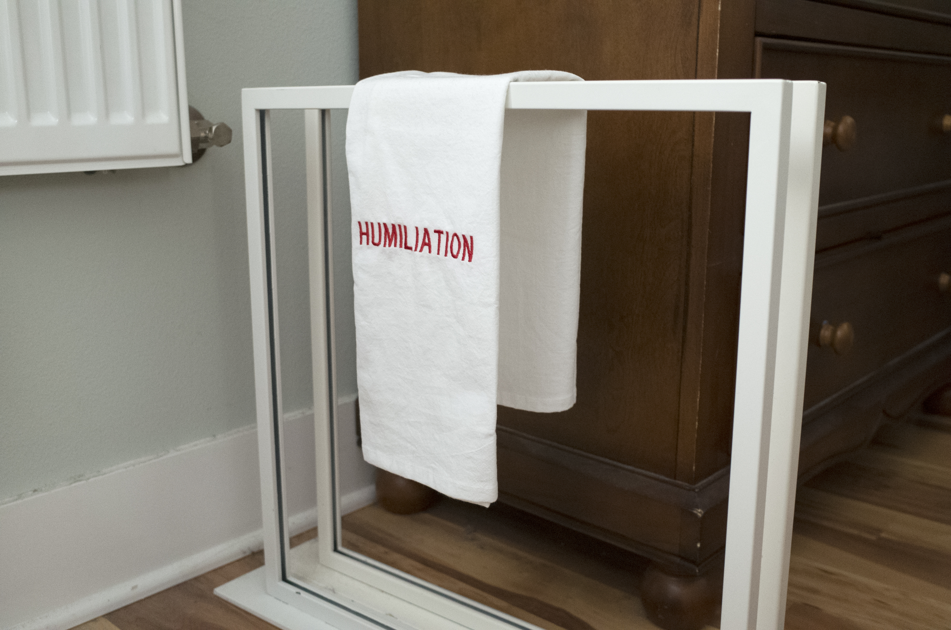 Two white metal frames stand upright and parallel to each other with a small gap in between on the floor of a domestic space. A folded white towel with the word ‘humiliation’ in red embroidery is draped over the top of the two frames.